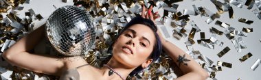 A young woman with short blue hair holds a disco ball in front of a bunch of confetti, creating a magical and festive scene.