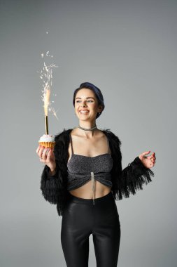 A young woman joyfully holding a cupcake with a sparkler, celebrating a birthday in a stylish setting.