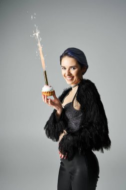A young woman with short, dyed hair posing in stylish attire, holding a cupcake with a sparkler.
