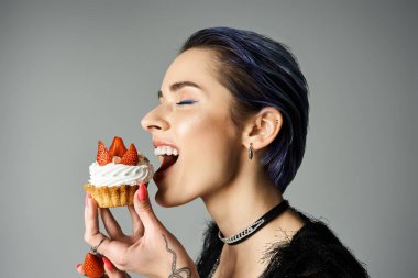 A young woman with short dyed hair smiling while eating a cupcake topped with strawberries. clipart