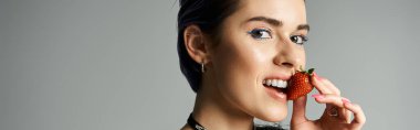 Stylish, young woman joyfully holds a ripe strawberry up to her lips in a vibrant studio setting.