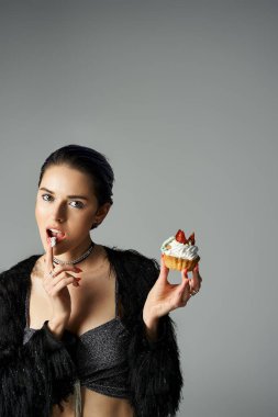 A stylish young woman with dyed hair poses with a cupcake, taking a bite from it.