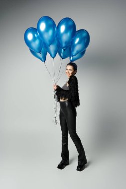 A young woman with short dyed hair and stylish attire joyfully holds a bunch of blue balloons.