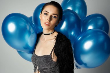 A stylish young woman with short dyed hair stands among a bunch of blue balloons in a studio setting.