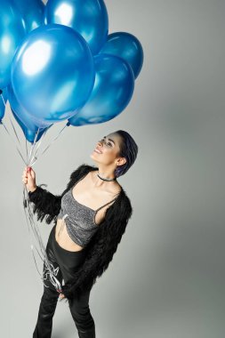 A stylish, young woman with short dyed hair poses gracefully while holding a bunch of vibrant blue balloons.