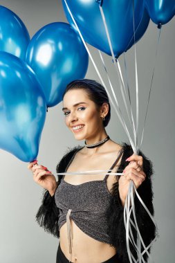 A stylish young woman with short dyed hair delightfully embraces a bunch of blue balloons in a studio setting.