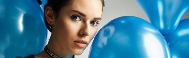 A stylish young woman with short dyed hair holds a collection of beautiful blue balloons in a studio setting.