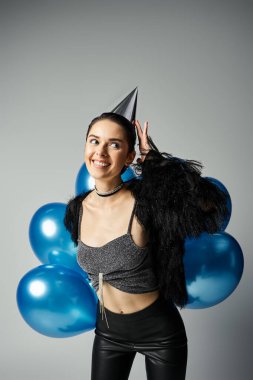 A stylish young woman with short dyed hair celebrates with a party hat and a bunch of colorful balloons.