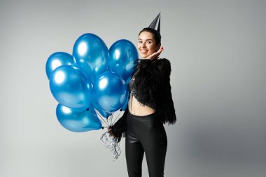 A stylish, young woman with colorful hair holding a cluster of vibrant blue balloons in a studio setting.