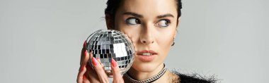 A young woman with short dyed hair poses in stylish attire, holding a disco ball in front of her face.