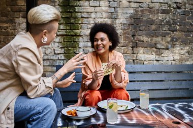 Diverse couple of lesbians enjoying a meal together at a cafe table. clipart