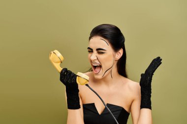 Pop sanat makyajlı esmer ve siyah elbiseli kadın canlı bir arkaplan karşısında sarı bir telefon tutuyor..