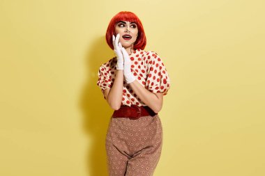 Popüler sanat makyajlı kızıl saçlı bir kadın sarı arka planda puantiyeli gömlek ve beyaz eldivenler giyiyor..