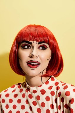 Parlak pop sanat makyajlı, sarı arka planda benekli bluz giyen çarpıcı kızıl saçlı bir kadın..