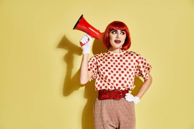 Yaratıcı pop sanat makyajlı kızıl saçlı güzel bir kadın sarı arka planda kırmızı ve beyaz bir megafon tutuyor..