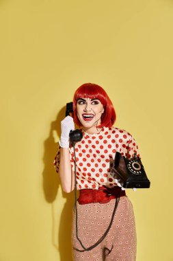 Puantiyeli güzel bir kızıl saçlı kadın yaratıcı pop sanat makyajıyla, canlı sarı bir arka planda telefon tutarken gülümsüyor..
