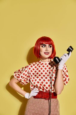 Canlı kızıl saçlı, yaratıcı pop sanat makyajı ve puantiyeli bluzu olan bir kadın sarı arka planda bir telefon tutuyor..