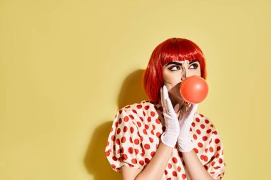 Popüler sanat makyajlı güzel bir kızıl kadın, benekli bluz, sarı arka planda balon şişiriyor..