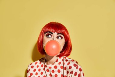 Palyaço burunlu kızıl saçlı bir kadın, pop sanat makyajı, puantiyeli bluz, sarı arka planda balon şişiriyor..