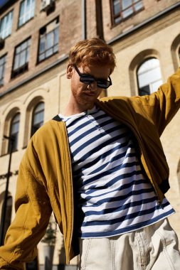 A young man with red hair strolls through the city in a debonair striped shirt and sunglasses. clipart