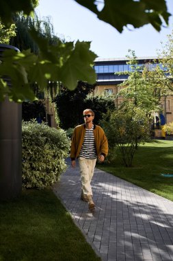 Şık giyimli, genç, kızıl saçlı bir adam şehir parkında kaldırımda yürüyor..