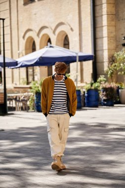 Genç kızıl saçlı bir adam bir binanın önünde yürüyor, güler yüzlü giyinmiş..