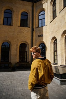 Kızıl saçlı güler yüzlü genç bir adam yüksek bir şehir binasının önünde güvenle duruyor..