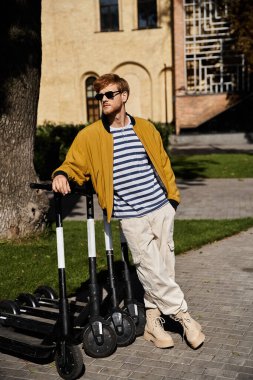 A young red-haired man in debonair attire standing next to black scooters in the city. clipart