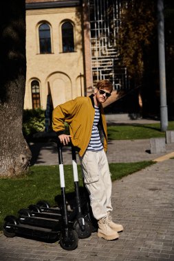 Young red-haired man in debonair attire standing beside scooters in the city. clipart