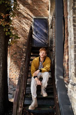 Genç, kızıl saçlı bir adam şık bir şehirde merdivenlerde zarif bir şekilde oturuyor..