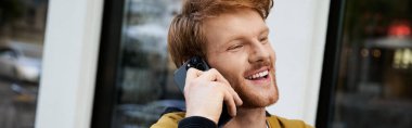 Şık, kızıl saçlı genç bir adam şehirde bir kafede otururken telefonla konuşuyor..