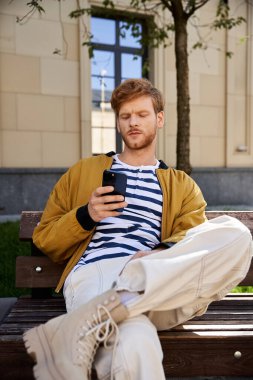 A stylish red-haired man sitting on a bench, using a cell phone. clipart