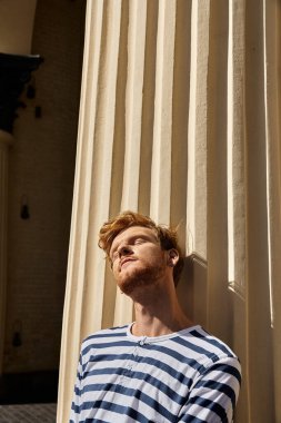 Kızıl saçlı şık bir genç adam zarif bir şekilde renkli bir perdenin yanında duruyor..