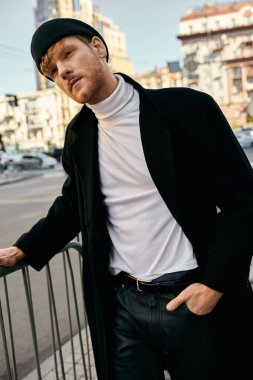 Şık giyinmiş kızıl saçlı genç bir adam elleri kalçasında çitlere yaslanıyor..