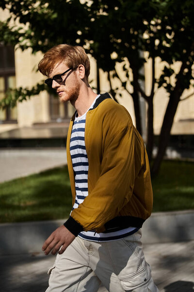 Молодой рыжий мужчина уверенно прогуливается по улице в коричневой куртке..