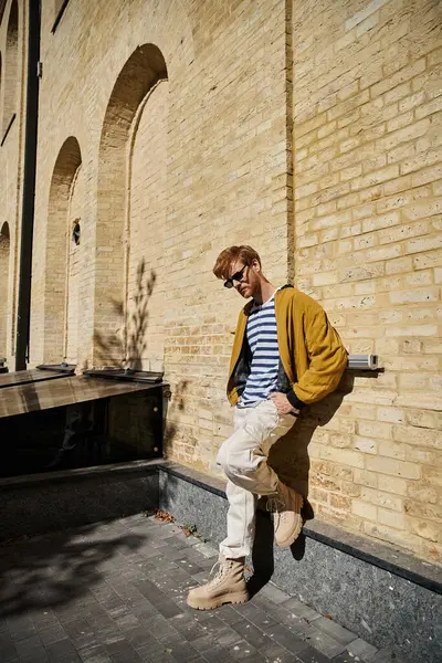 stock image A young man with red hair leans against a brick wall in a city setting, exuding debonair charm.