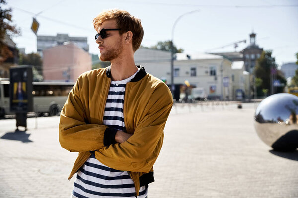 Young man with red hair and debonair attire, arms crossed, standing in middle of street.