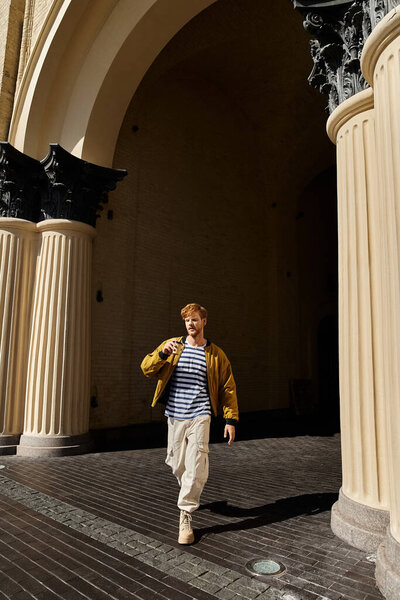 A young red-haired man in debonair attire walking down a city street flanked by tall pillars.