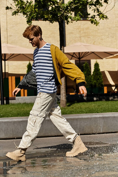 Стильный молодой человек с рыжими волосами пересекает городскую улицу рядом с фонтаном.