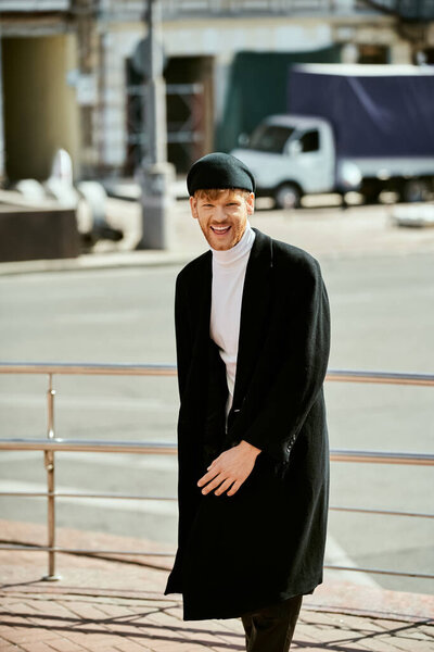 Молодой человек с рыжими волосами, в элегантном черном пальто и шляпе, улыбается.