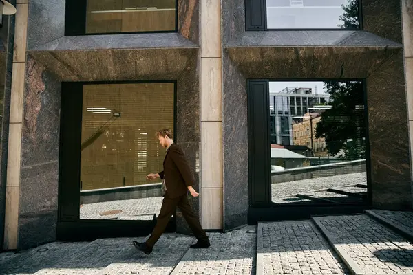 stock image Young red-haired man in debonair attire walking past a tall building in the city.