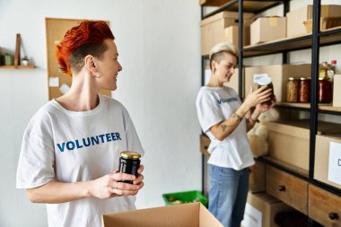 Young lesbian couple in volunteer t-shirts organizing boxes and cans for charity work in the room. clipart