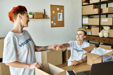 A woman in a volunteer shirt holding hands with her loved one amidst boxes in a crowded room. clipart