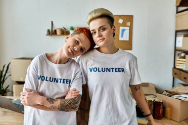 Two young women, wearing volunteer t-shirts, stand side by side in a room, exuding unity and dedication to charity work. clipart