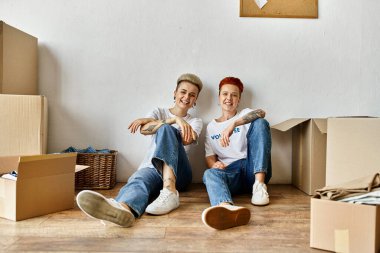 Two women, volunteers in charity shirts, sit amidst moving boxes, sharing a moment of connection and unity. clipart