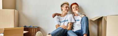 A young lesbian couple in volunteer t-shirts sitting on cardboard boxes, united in charity work. clipart