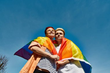 LGBTQ topluluğunun bir parçası olan iki kadın, açık mavi gökyüzünün altında gökkuşağı bayrağıyla birlikte duruyorlar..