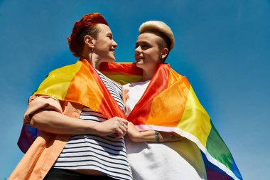 LGBTQ topluluğunun bir parçası olan iki genç kadın, gökkuşağı bayrağıyla mavi bir gökyüzünün altında birlikte duruyorlar..