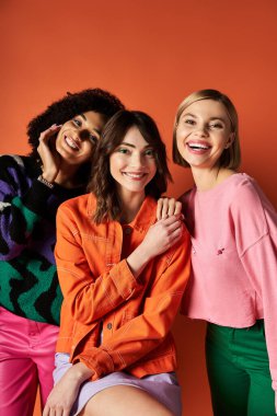 Three beautiful multicultural women posing in stylish clothes against a striking orange backdrop. clipart