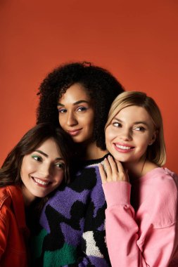 Farklı kültürlerden üç genç ve şık kadın çarpıcı bir turuncu zemine karşı birlikte poz veriyorlar..